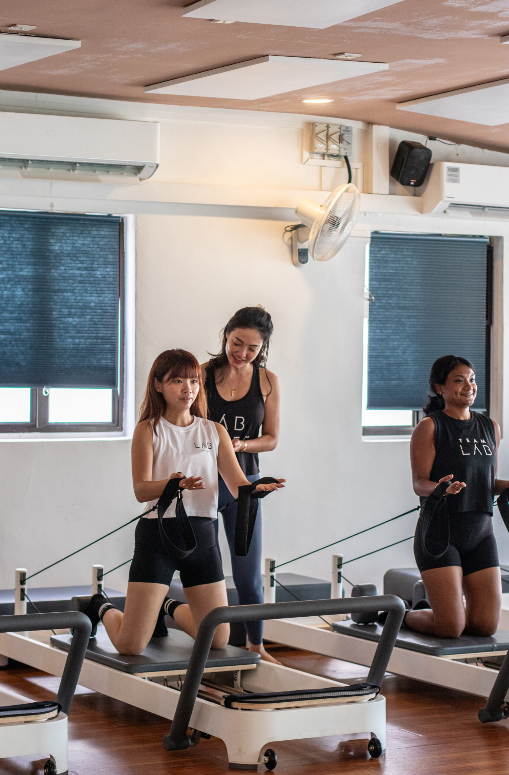 A small group in Lab Studios, smiling and working with a instructor who is offering a gentle correction on a Reformer.